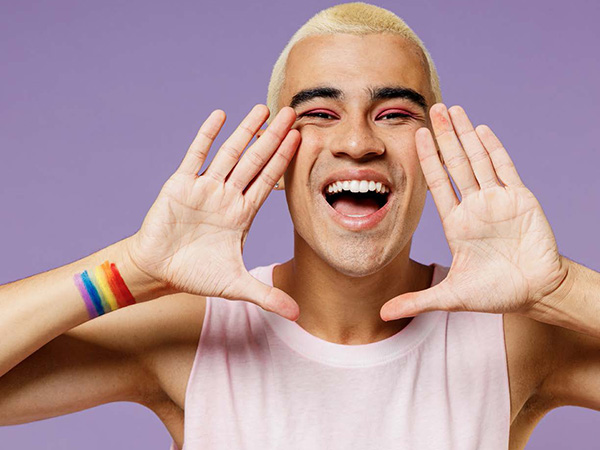 Image of an individual smiling and yelling with a pride flag tattoo standing in front of a light purple background.