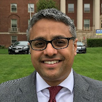 Indian-American Science Policy Branch Chief Sandeep Dayal, Ph.D., standing in front of an NIH campus building, several cars, and a grassy area.