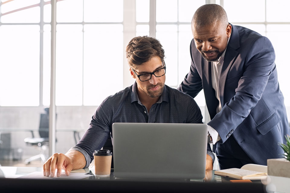 A Black supervisor stands behind his seated employee, pointing to something unseen on a laptop before them.