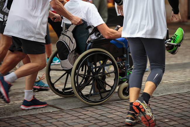 A group of people jogging. One of the runners is pushing a man in a wheelchair.