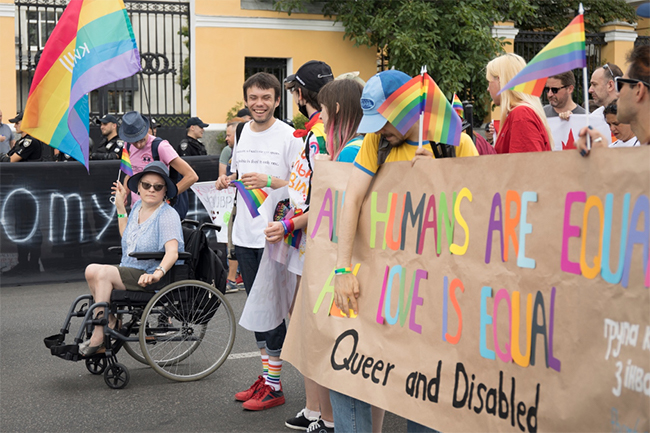 Group of people at PRIDE parade holding up banner that says: All Humans Are Equal; All Love is Equal: Queer and Disabled