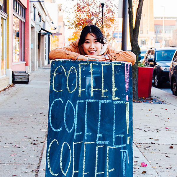 Olive Jung leaning on a chalkboard sign with coffee written on it.