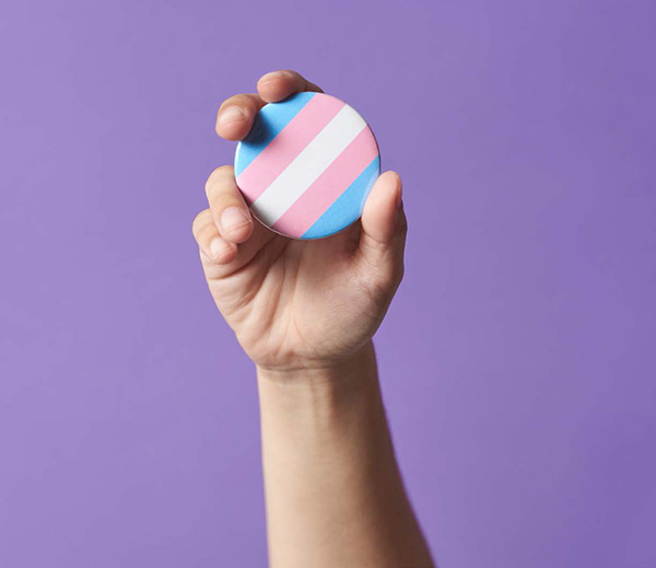 A hand holding up a transgender flag button.