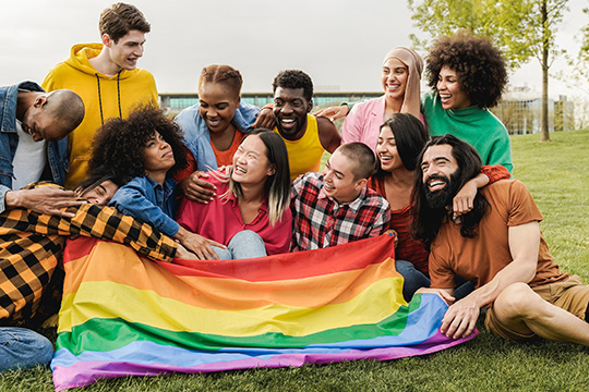 A diverse group of friends hold a pride flag while smiling and having fun outdoors.