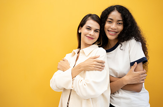 Two women show self-love by hugging themselves in front of a bright yellow background.