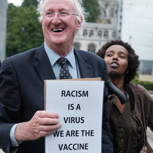 A white male smiling holding a sign that reads Racism in a virus we are the cure with a black female behind him