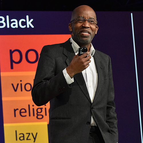 A black man with glasses and wearing a black suit giving a speech