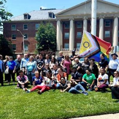 A group photo of Salutaris members in front of NIH Building 1 with the Progress Pride flag behind the group. 