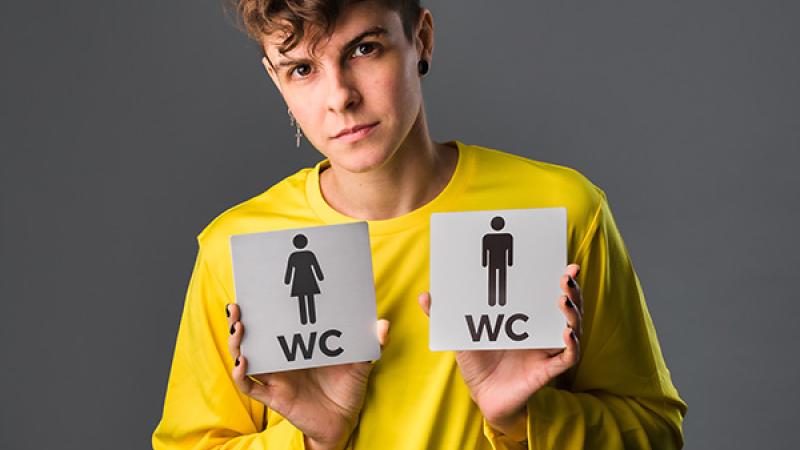 Image of a person in a yellow shirt holding two gendered bathroom signs.