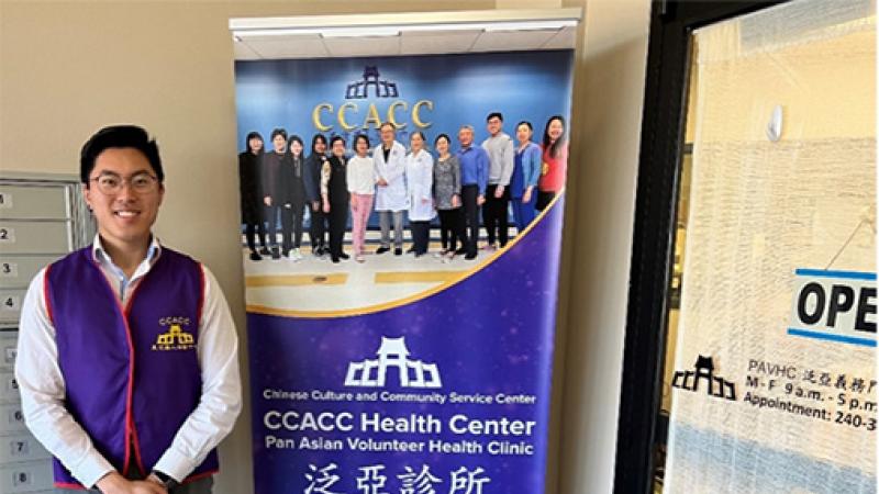 Author and IRTA Fellow, Simon Liu wearing a blue vest and standing in front of a Chinese Culture and Community Service Center banner.