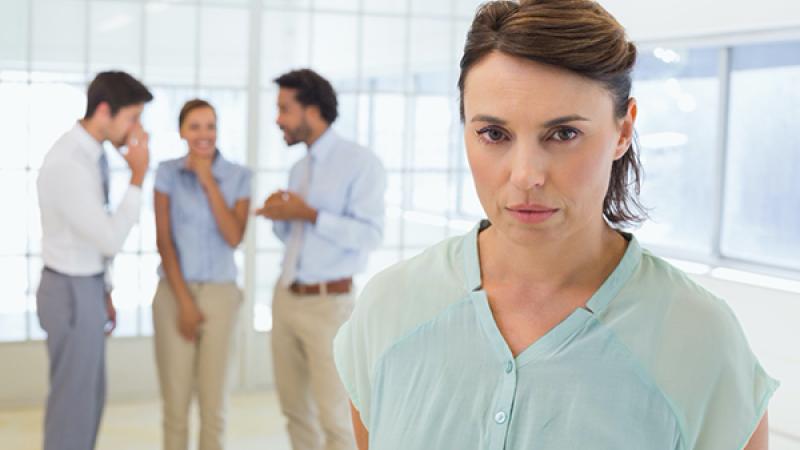 A sad businesswoman standing in front of a group of colleagues who are gossiping about her at the office.