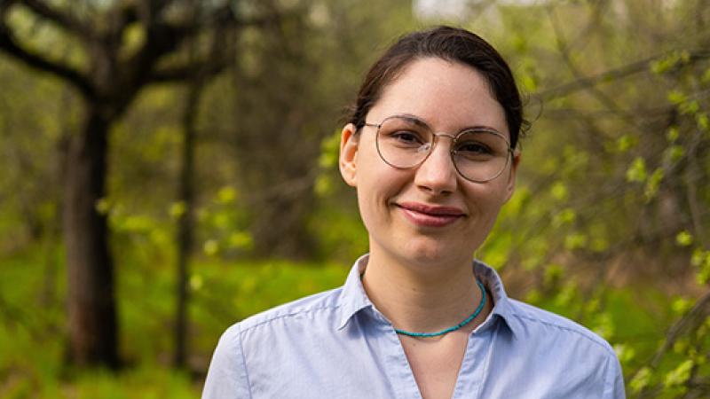 A photo of a woman with glasses looking at the camera with trees in the background.