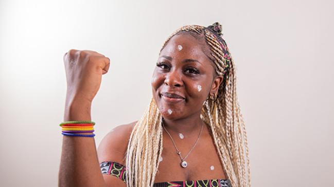 A Black lesbian woman with her arm raised shows her bicep and wears colorful, rainbow colored bracelets. She wears an African print halter top with black, green, purple, and pink geometric shapes.