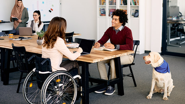 A man with a canine service dog meets with a woman using a wheelchair in a diverse office.  